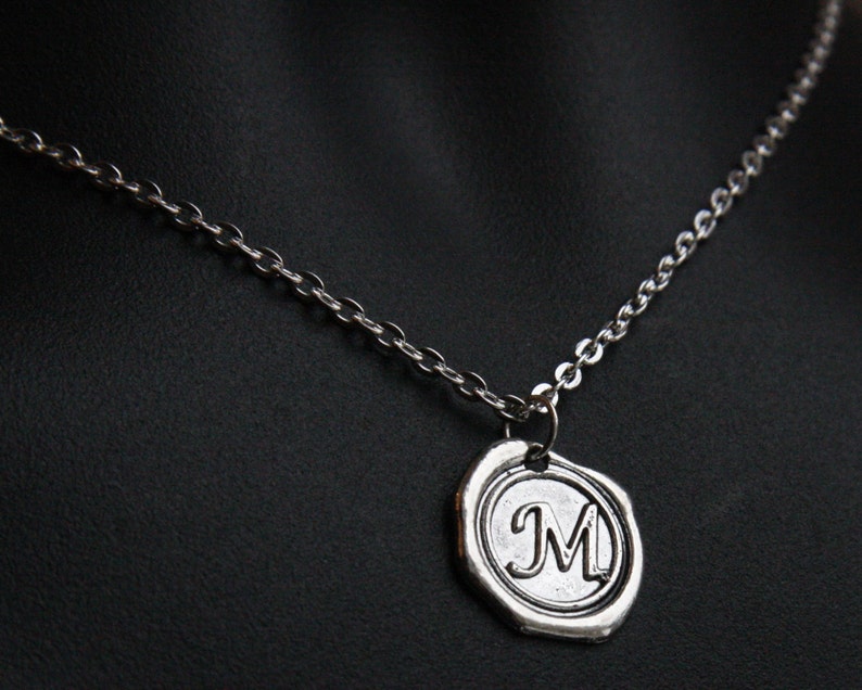 May include: Silver-toned necklace featuring a pendant with the letter "M" inside a circular frame. The pendant has a textured, antique appearance, and hangs from a delicate chain. The necklace is displayed against a dark background.