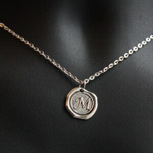 May include: Silver-toned necklace featuring a pendant with the letter "M" inside a circular, embossed design. The pendant hangs from a delicate chain, displayed against a black background.
