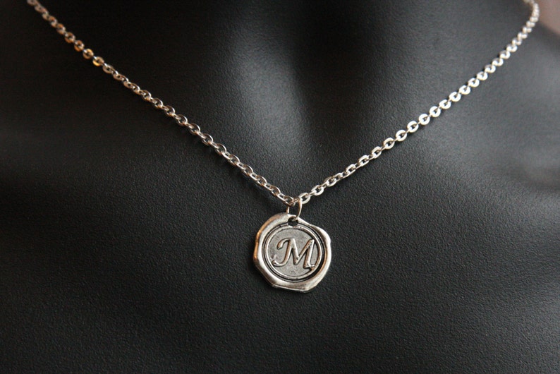 May include: Silver-toned necklace featuring a pendant with the letter "M" inside a circular, embossed design. The pendant hangs from a delicate chain, displayed against a black background.