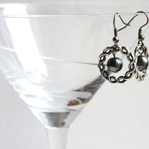 May include: A pair of silver earrings with a black pearl in the center of each earring. The earrings are in the shape of a circle with a braided design.