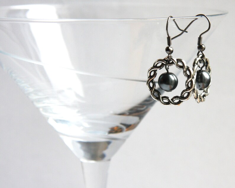 May include: A pair of silver earrings with a black pearl in the center of each earring. The earrings are in the shape of a circle with a braided design.