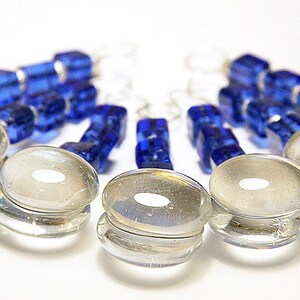 May include: Clear glass bottle stoppers with a circular design and a small white center. The stoppers are arranged in a semi-circle, with blue glass bead keychains in the background. The image is set against a white background.
