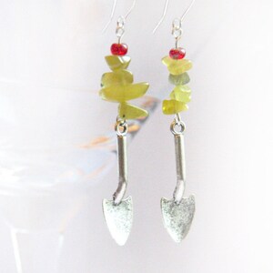 May include: Silver earrings with a green stone and red bead dangle, shaped like a small shovel.