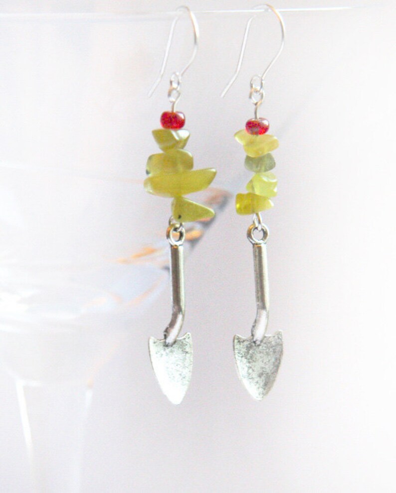 May include: Silver earrings with a green stone and red bead dangle, shaped like a small shovel.