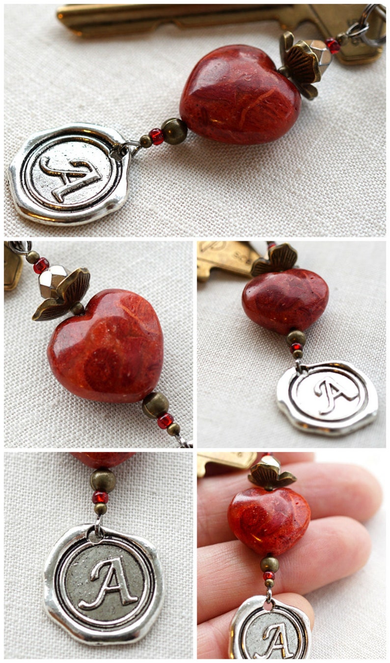 May include: A silver charm with the letter "A" in a wax seal design, attached to a red coral heart charm and a silver key charm. The charms are strung on a chain with red and silver beads.