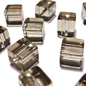 May include: Close-up of several clear, square-shaped beads with a smoky gray hue. Each bead has a hole running through the center, and some have horizontal lines etched into their surfaces. The beads are scattered on a white surface.