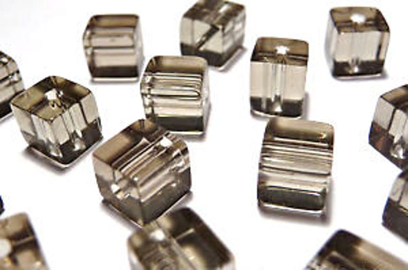May include: Close-up of several clear, square-shaped beads with a smoky gray hue. Each bead has a hole running through the center, and some have horizontal lines etched into their surfaces. The beads are scattered on a white surface.