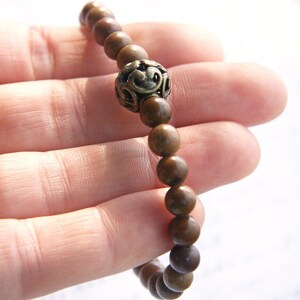 May include: A brown wooden beaded bracelet with a decorative metal charm. The bracelet is made of round beads and has a single metal charm with a floral design.