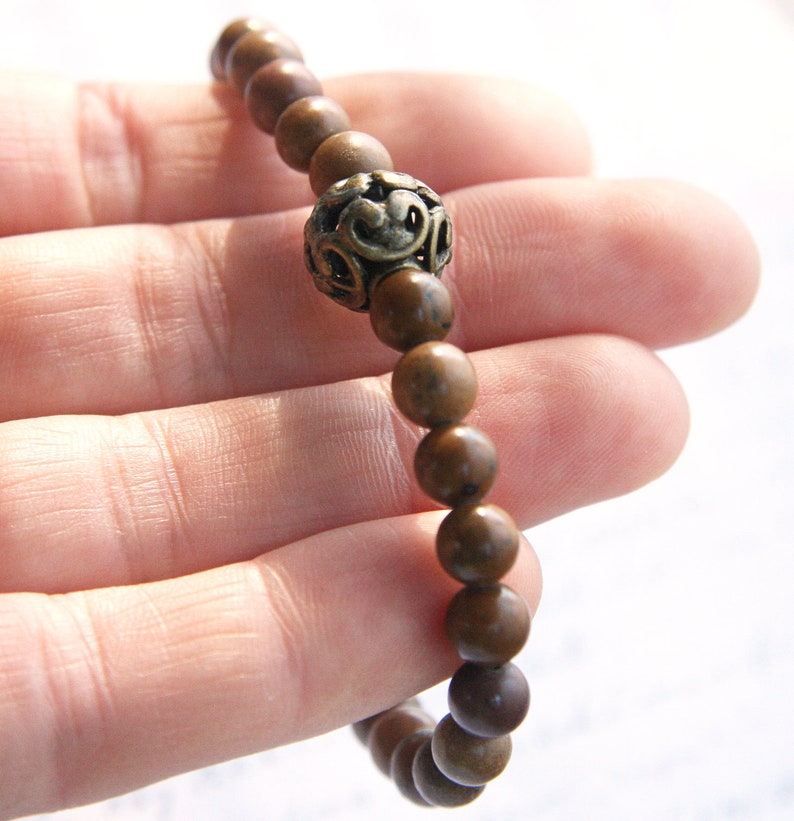 May include: A brown wooden beaded bracelet with a decorative metal charm. The bracelet is made of round beads and has a single metal charm with a floral design.