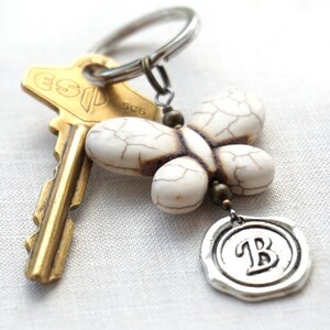 May include: White butterfly-shaped charm with brown veins, attached to a silver key ring with a gold key and a silver charm with the letter 'B' on it.