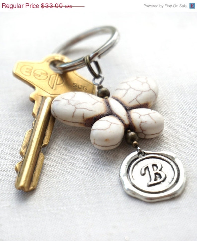 May include: White butterfly-shaped charm with brown veins, attached to a silver key ring with a gold key and a silver charm with the letter 'B' on it.