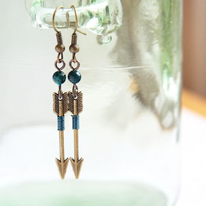 May include: A pair of gold arrow earrings with turquoise beads. The earrings are hanging from a glass jar.