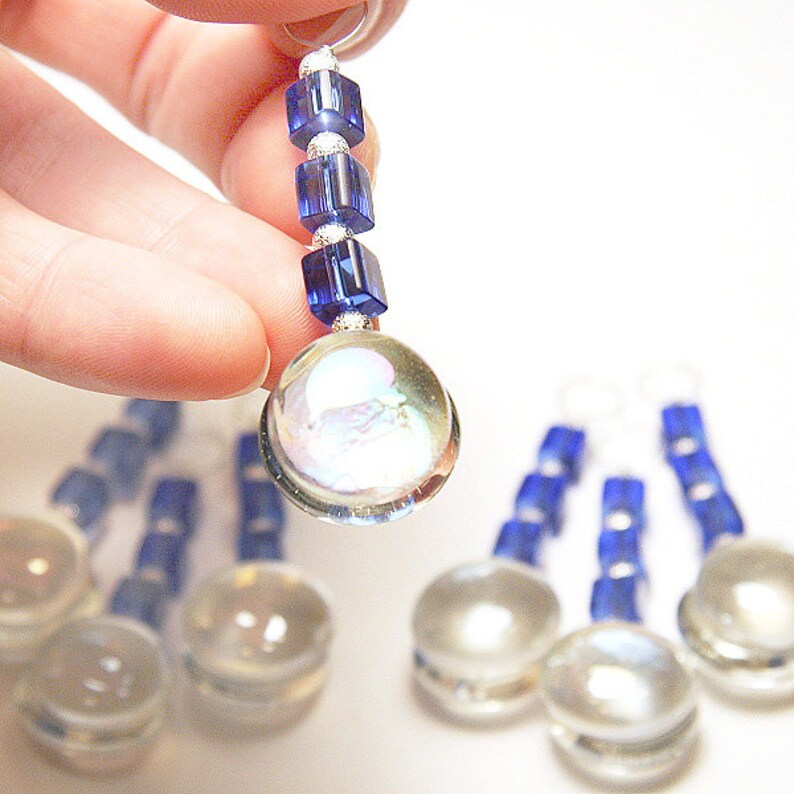 May include: Close-up of a handmade earring with a clear, iridescent glass orb and a blue beaded accent. The earring is suspended from a silver-colored ring. Additional earrings are visible in the background.
