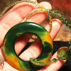May include: A green and orange agate stone pendant with a ring in the center, hanging from a gold and green beaded necklace.