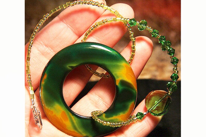 May include: A green and orange agate stone pendant with a ring in the center, hanging from a gold and green beaded necklace.