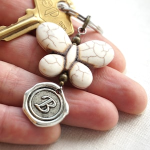 May include: White butterfly charm with a brown wax seal charm with the letter "B" on a gold key chain.