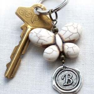 May include: A white butterfly charm with a silver key ring and a gold key. The butterfly charm has a brown wax seal with the letter "B" on it.