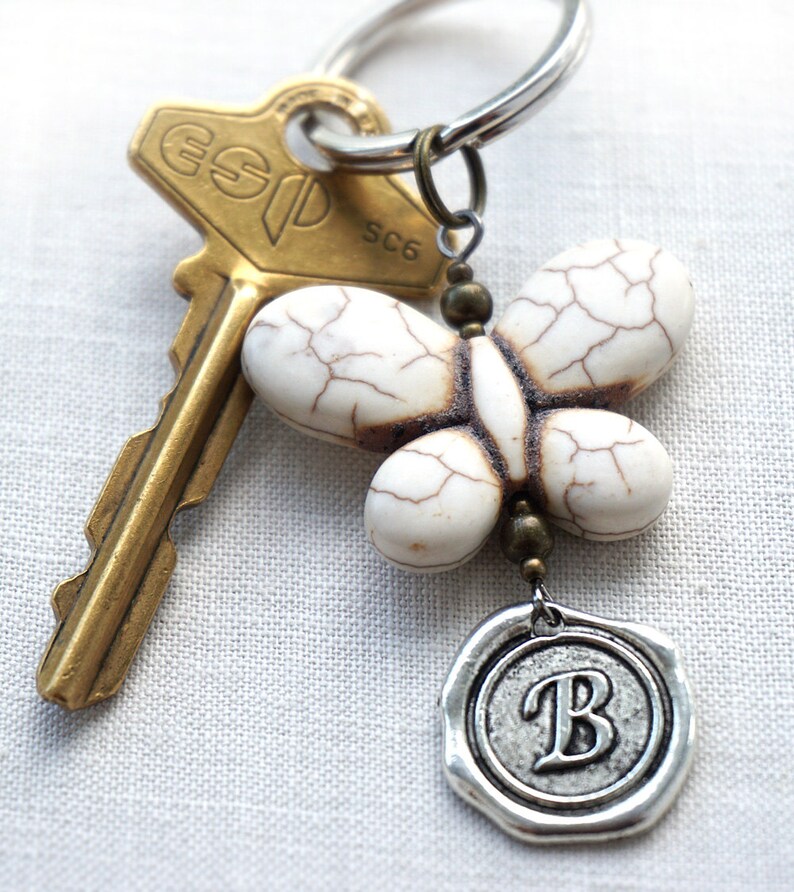 May include: A white butterfly charm with a silver key ring and a gold key. The butterfly charm has a brown wax seal with the letter "B" on it.