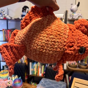 Könnte beinhalten: Handgefertigter, orange- und pfirsichfarbener Häkelgoldfisch mit großen, schwarzen Augen. Das Plüschtier hat detaillierte Flossen und ein strukturiertes, weiches Aussehen, perfekt für ein Kinderzimmer oder als einzigartiges Dekorationsstück.