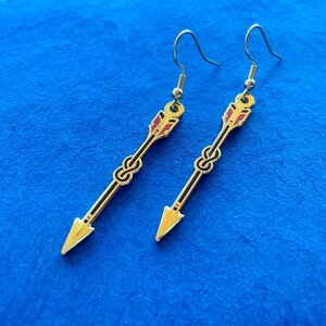 May include: A pair of gold-colored arrow earrings with black and red accents. The earrings feature a knot design in the center and a hook closure. The arrowheads are a bright gold color.