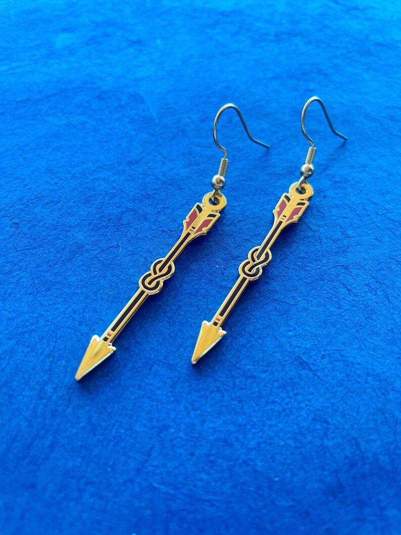 May include: A pair of gold-colored arrow earrings with black and red accents. The earrings feature a knot design in the center and a hook closure. The arrowheads are a bright gold color.