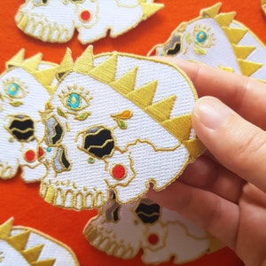 May include: White embroidered skull patch with a gold crown, detailed with black, red, blue and green accents. The skull has a third eye with a blue iris and a gold rim.