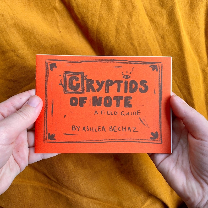 May include: A small orange booklet with the title "Cryptids of Note: A Field Guide by Ashlea BeChaz" printed in black.