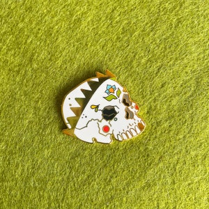 May include: White enamel pin of a skull wearing a crown with colorful flowers. The skull has black eye sockets and a red dot on the forehead.