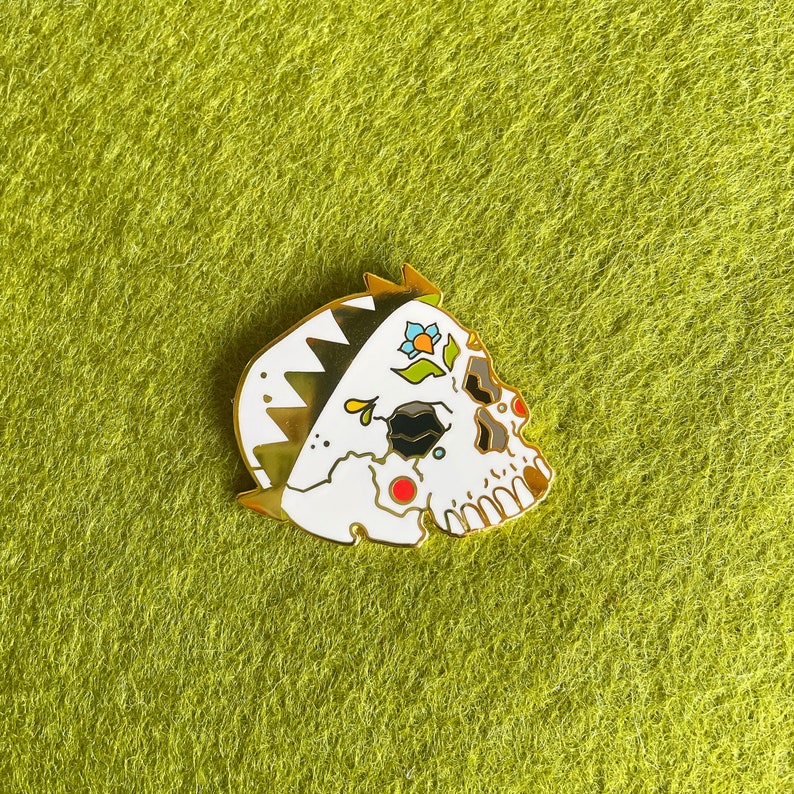 May include: White enamel pin of a skull wearing a crown with colorful flowers. The skull has black eye sockets and a red dot on the forehead.
