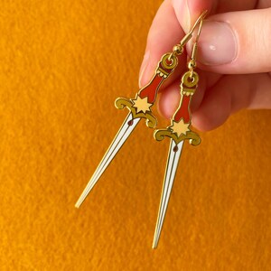 May include: Gold tone earrings with a red and white dagger design. The daggers have a star and a red drop of blood on them.