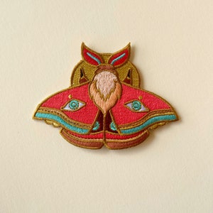 Op de afbeelding: Gestikte patch van een roze nachtvlinder met gouden en turquoise accenten. De vlinder heeft grote ogen en een gedetailleerd ontwerp.