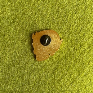 May include: Gold-colored enamel pin with a glitter finish. The pin features a cartoon-style character with a large head and a small body. The pin has a black metal backing with a safety clasp.