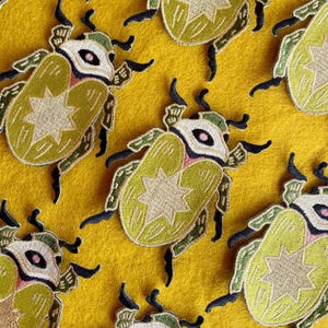 May include: A set of six embroidered iron-on patches featuring green and yellow beetles with a white star on their backs. The beetles have a pink and white eye design.