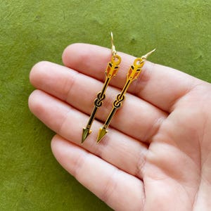 May include: Gold-colored arrow earrings with black and orange details. The earrings have a hook closure and a knot design. The arrowheads are a metallic gold color. The earrings are displayed on a green background.