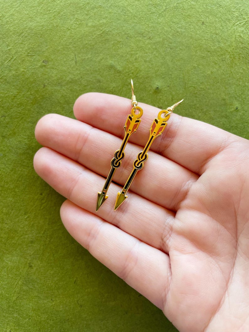 May include: Gold-colored arrow earrings with black and orange details. The earrings have a hook closure and a knot design. The arrowheads are a metallic gold color. The earrings are displayed on a green background.