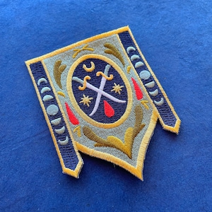 May include: Embroidered patch in a shield shape with a navy blue border and gold accents. The center features crossed swords, stars, and a crescent moon design. The patch has a light blue and gold background with red accents.