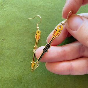 May include: Gold-colored arrow earrings with black and red accents. The earrings feature a hook closure and a detailed arrow design, with a knot detail in the center. The earrings are held against a green background.