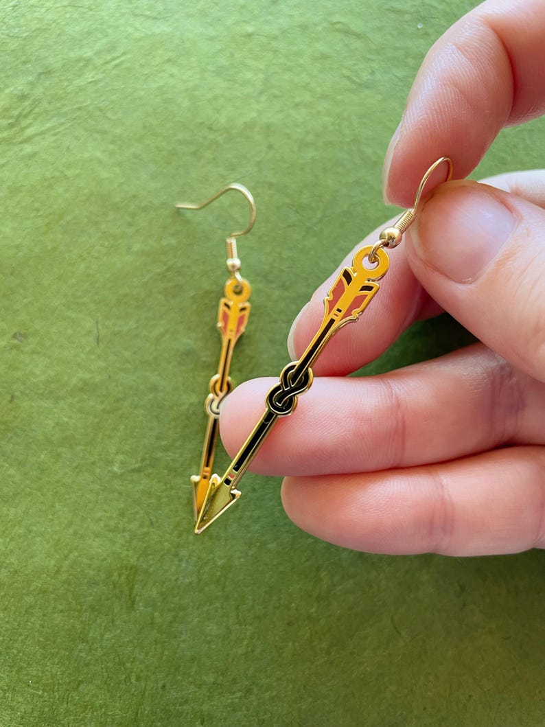 May include: Gold-colored arrow earrings with black and red accents. The earrings feature a hook closure and a detailed arrow design, with a knot detail in the center. The earrings are held against a green background.