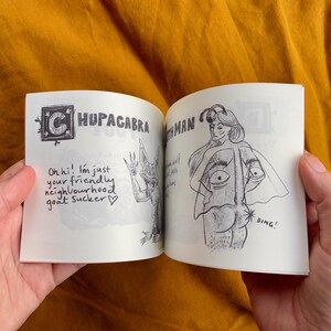 May include: An open book with illustrations of a chupacabra and a mothman. The chupacabra is a creature with large ears and sharp teeth. The mothman is a humanoid figure with large wings. The text on the page reads "Oh hi! I'm just your friendly neighbourhood goat sucker." and "Mothman".