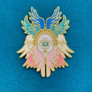 May include: Embroidered patch with a gold halo and a single blue eye, surrounded by pink and white wings with gold accents.