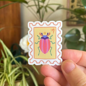 Puede incluir: Una pegatina blanca con un borde amarillo y naranja, que presenta un escarabajo rosa y azul con patas negras y un círculo amarillo en la espalda. La pegatina tiene un borde blanco con un borde festoneado y el texto "寿" en rojo.