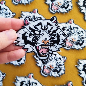 May include: A white embroidered patch depicting a tiger with a third eye in the centre of its forehead. The tiger has a black and white striped pattern and an open mouth with pink teeth. The patch is on a yellow felt background.
