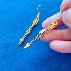 May include: A pair of gold-colored arrow-shaped earrings with black and red accents. The earrings have a hook closure and a pointed tip. The design features a decorative knot detail near the center of the arrow.