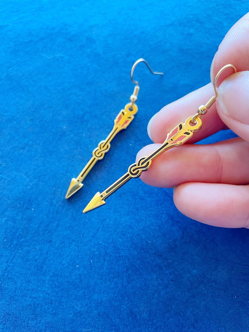 May include: A pair of gold-colored arrow-shaped earrings with black and red accents. The earrings have a hook closure and a pointed tip. The design features a decorative knot detail near the center of the arrow.