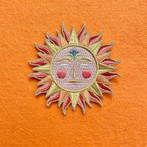 May include: Embroidered iron-on patch featuring a stylized sun with a smiling face and a third eye. The sun has a pink face, yellow rays, and red flames.
