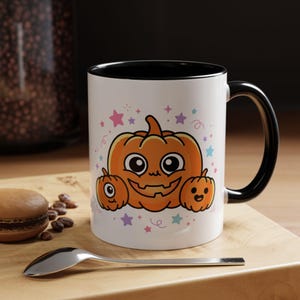May include: A white ceramic mug with a black interior and handle, featuring a graphic of three orange pumpkins with smiling faces surrounded by small colorful stars and swirls. The central pumpkin is large, flanked by two smaller pumpkins.