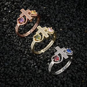 Puede incluir: Tres anillos con diseños de cruz y piedras preciosas. Los anillos son de oro, plata y oro rosa. Cada anillo tiene una cruz con piedras claras y una piedra preciosa a cada lado. Las bandas de los anillos tienen texto.