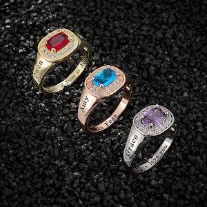 Puede incluir: Tres anillos personalizados con piedras rectangulares rojas, azules y moradas. Los anillos son dorados, oro rosa y plateados, cada uno con un halo de piedras más pequeñas. Cada anillo tiene un nombre y una frase grabados.