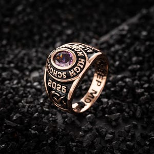 May include: A gold-colored class ring with a purple gemstone center. The ring features the words "HIGH SCHOOL" and "2025" in black lettering. The ring is set against a dark, textured background.