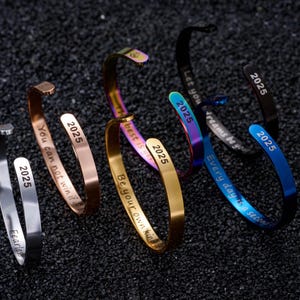 May include: Several open cuff bracelets in silver, gold, and iridescent finishes. Each bracelet is engraved with motivational phrases and the year "2025".